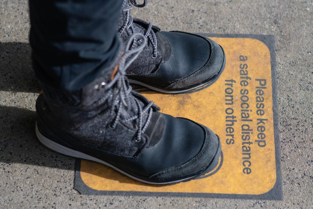 Black boots standing on a social distancing sign on pavement, emphasizing pandemic safety.